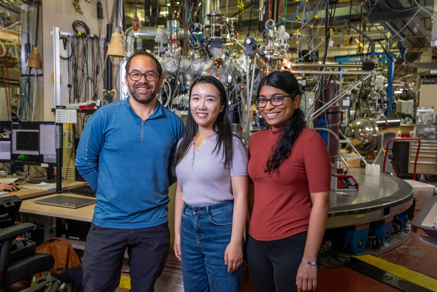 Three researchers stand in front of a complex experimental facility. The experiment consists of various colorful wires and pipes.