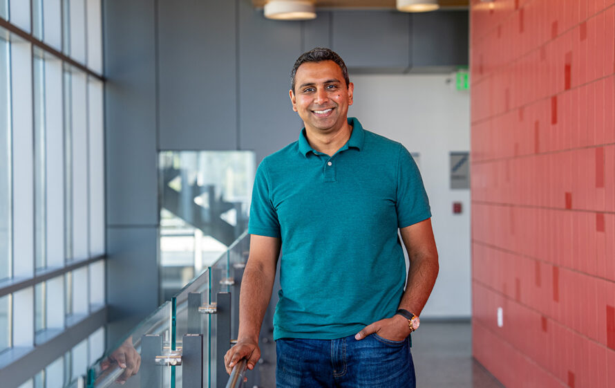 Researcher dressed in a teal-colored polo shirt and blue jeans posing near a stairway, between a red-colored wall and windows.