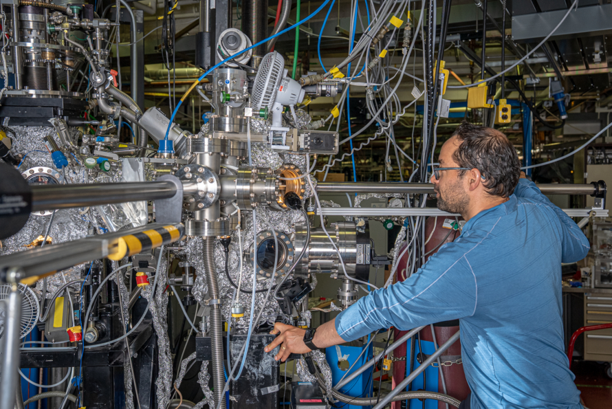 A researcher in a blue shirt and glasses stands to the right of a complex experimental setup. The setup is surrounded by wires and other hook-ups.