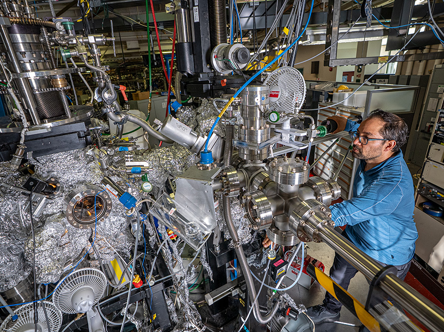 A researcher in a blue shirt and glasses stands to the right of a complex experimental setup. The setup is surrounded by wires and other hook-ups.