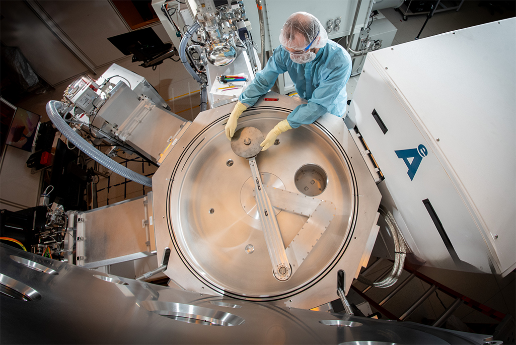 A scientist in light blue personal protective equipment adjusts a metallic disk inside the cluster tool – seen from above.