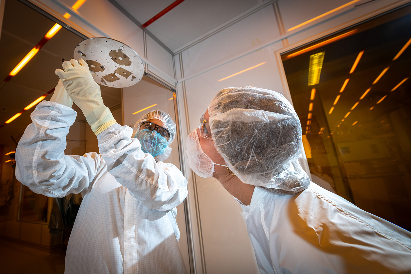 One researcher holds up a disk while another researcher peers at the underside. Both researchers are wearing white personal protective equipment and stand in front of orange reflective lab windows.