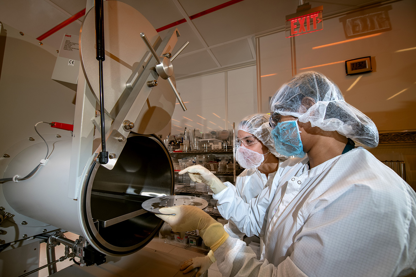 wo researchers in white personal protective equipment load a metallic, disk-like sample into a large tube on the cluster tool.