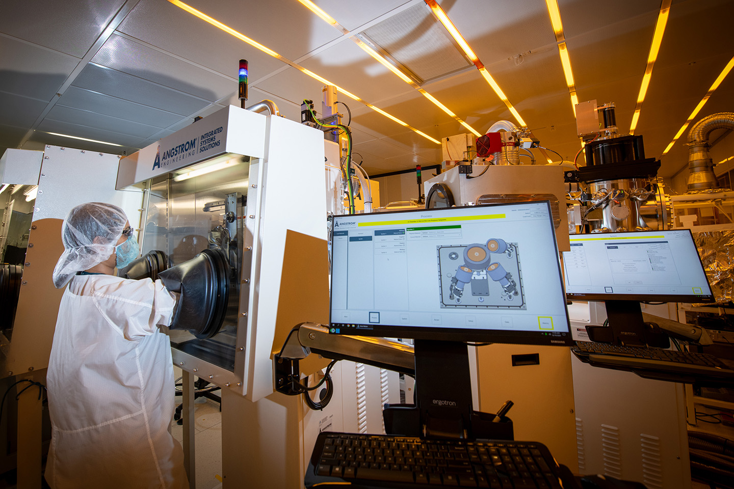 A researcher in white personal protective equipment stands with their hands in the gloves of an experimental hood. There are various monitors and instruments in an orange-tinged laboratory.