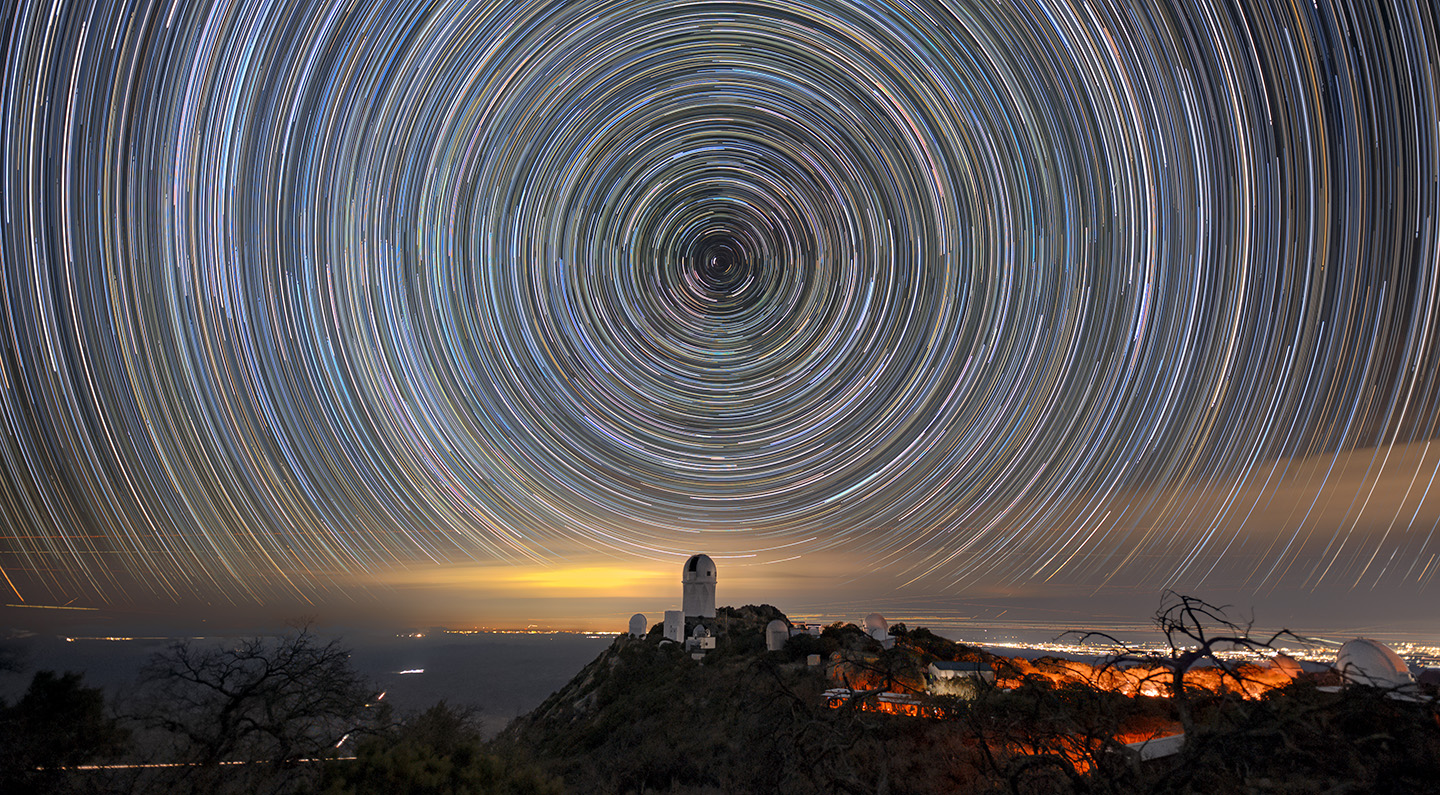 Circles of light on the night sky. A telescope dome atop a mountain is below the center of the circle.