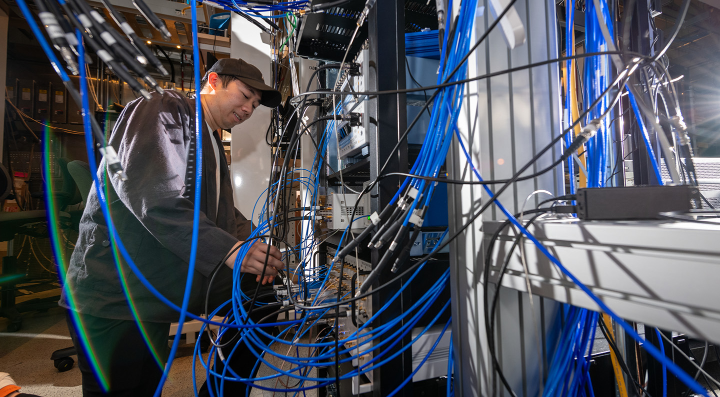 Researcher in a lab with blue-colored wires suspended against a large, gray metallic device.