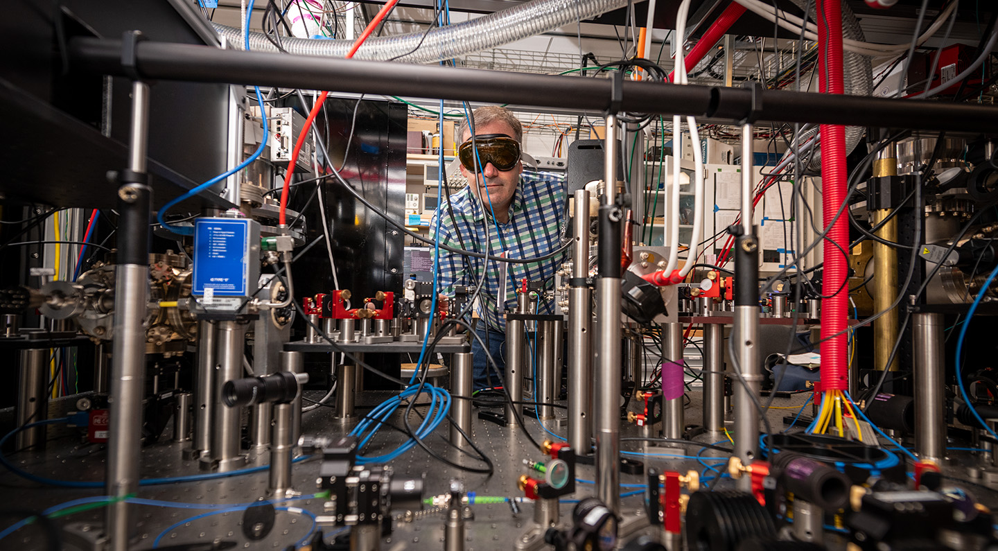 A researcher in a blue plaid shirt and black safety goggles stands behind a large laser experiment set up. The subject is blocked from colorful hanging wires and other apparatus.