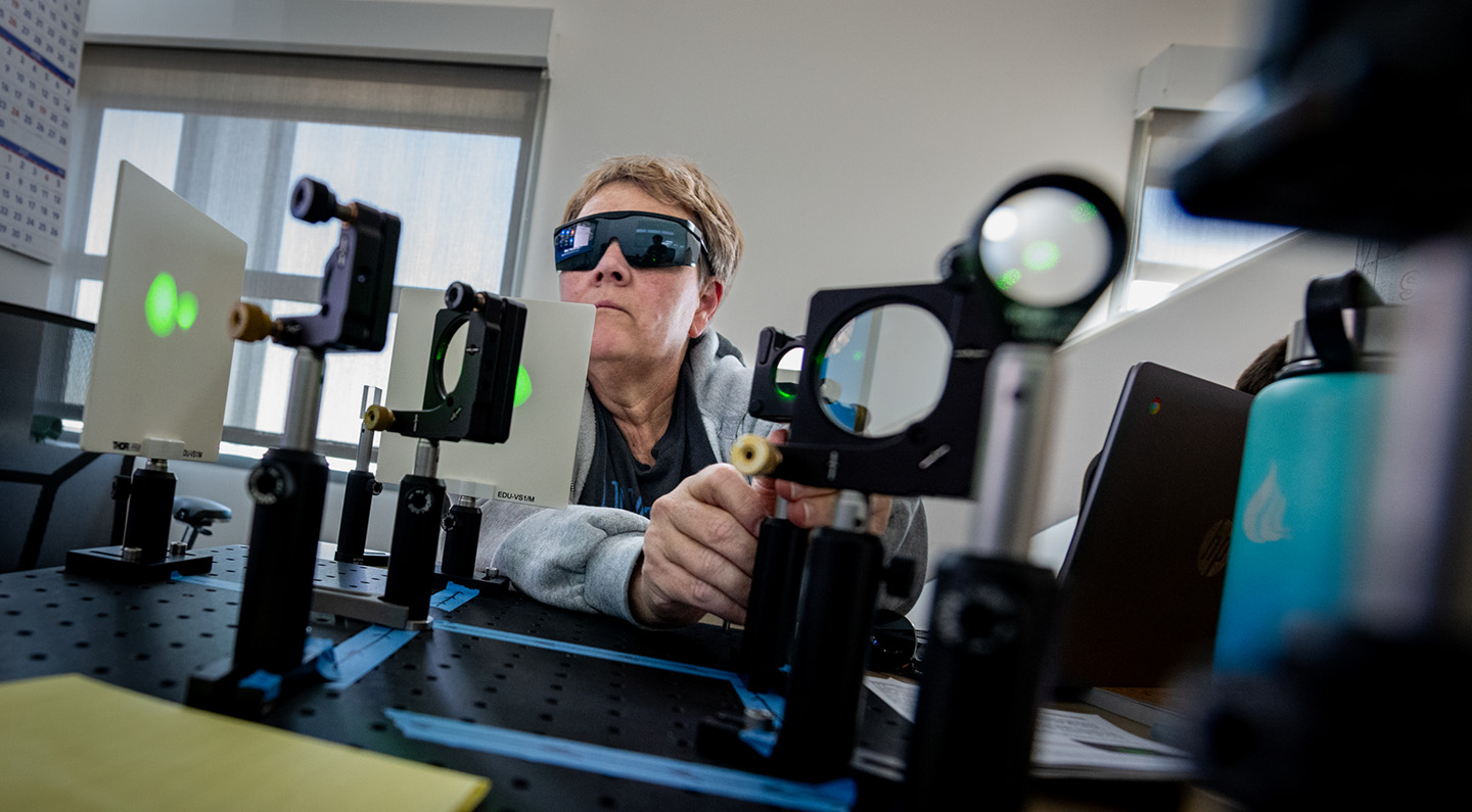 A teacher in protective glasses sets up an experiment on a table. The table displays multiple lens aparati.