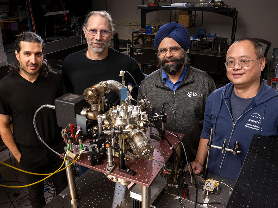 Four researchers in a dark lab space stand around a device with colorful wires and metallic pipes.