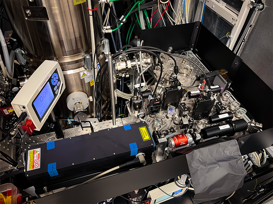 A close-up photograph of an intricate instrument. On the left is a thick metallic column with a small digital screen. In the foreground, a long black laser component connects to a complex open compartment filled with a dense array of mirrors, lenses, and specialized electronic parts, all surrounded by tightly bundled wires.