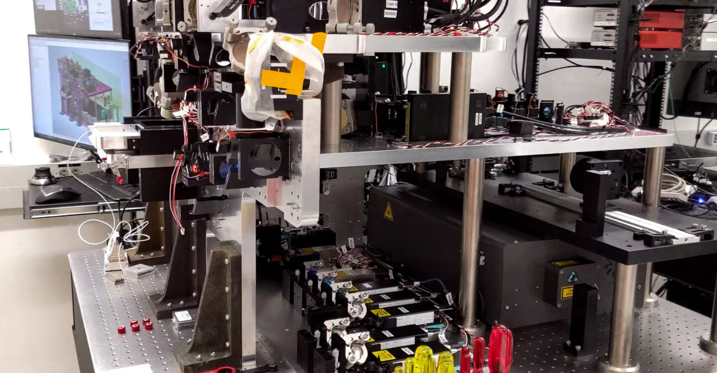 A complex laboratory assembly set on a metal breadboard table, filled with lenses, laser components, mirrors, and organized wiring. In the background to the left, a computer monitor displays a 3D computer-aided design rendering of the optical system.