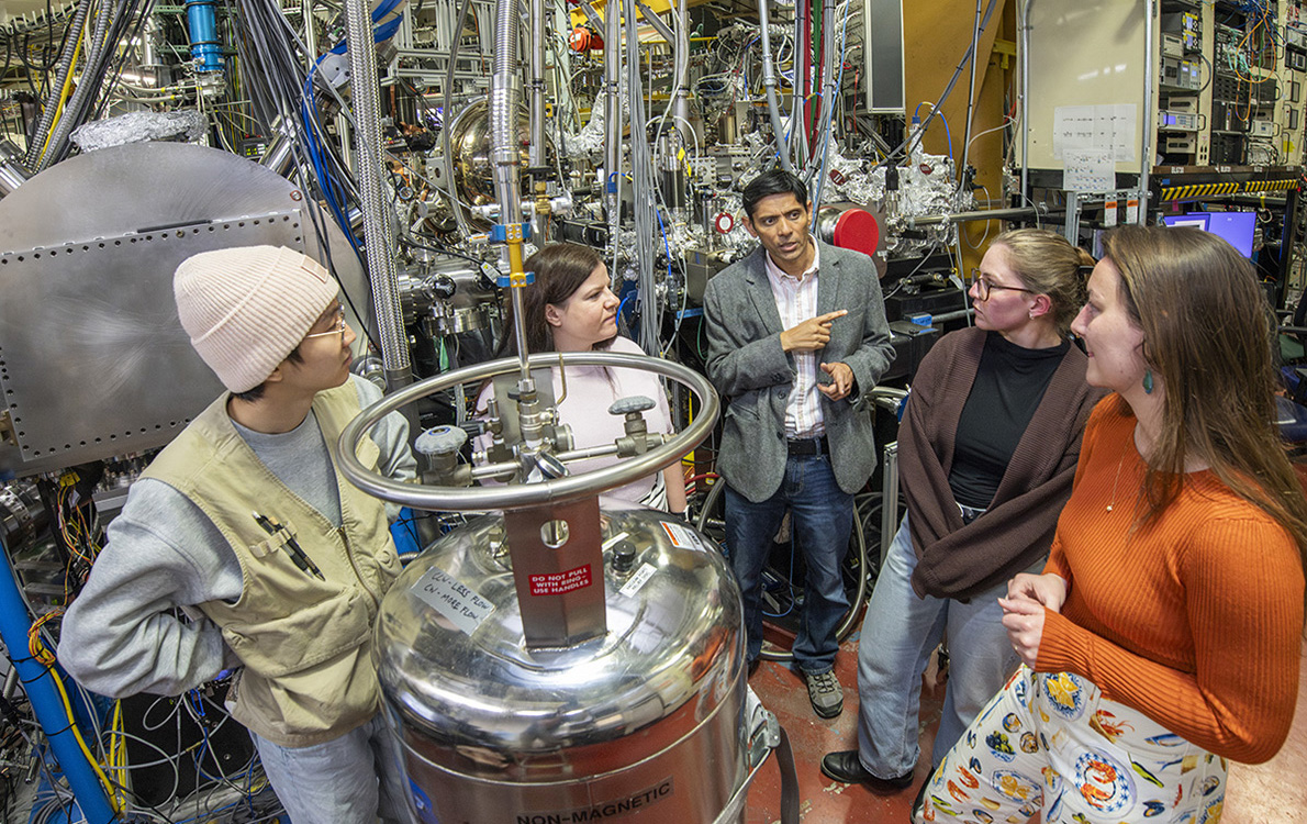 Five researchers meeting in a lab with large metallic instruments.