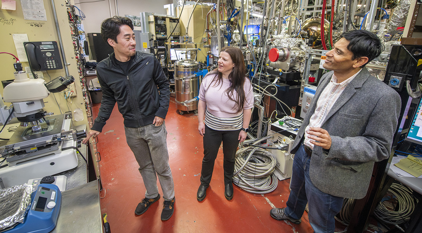 Three researchers meeting in a lab with large metallic instruments and an X-ray microscope.