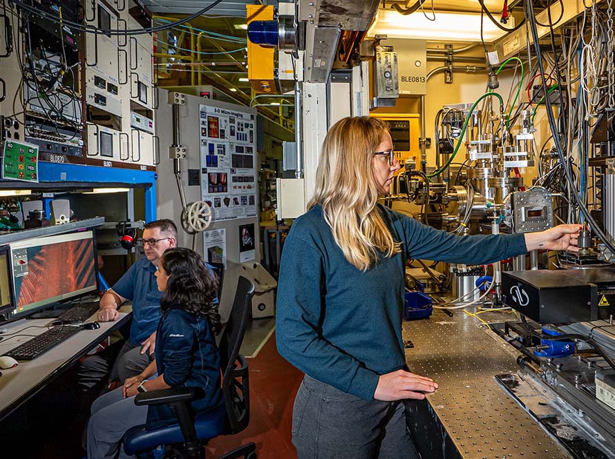 A female researcher wearing teal and gray-colored office clothes adjusting an instrument at the Advanced Light Source microtomography beamline. Two additional scientists are seen in the background looking at computer screens displaying microscopy imagery.