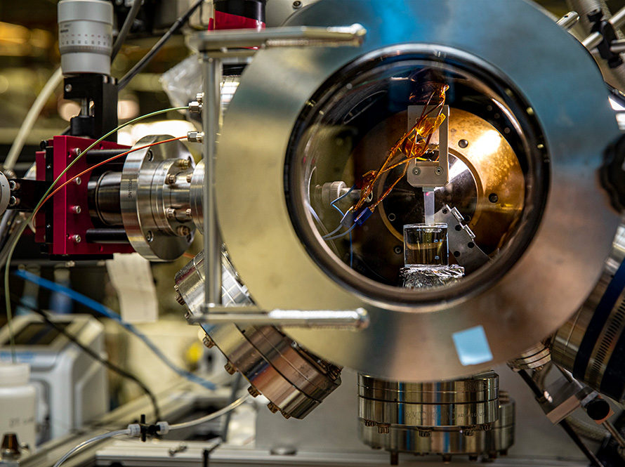 A small container and wires are seen through a round viewing chamber of a chrome instrument at the Advanced Light Source.