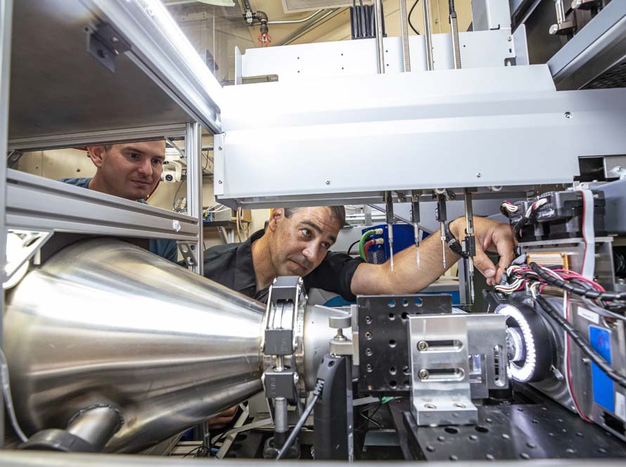 Two men are seen through gaps in a large chrome-colored instrument. One is reaching through and making small adjustments.