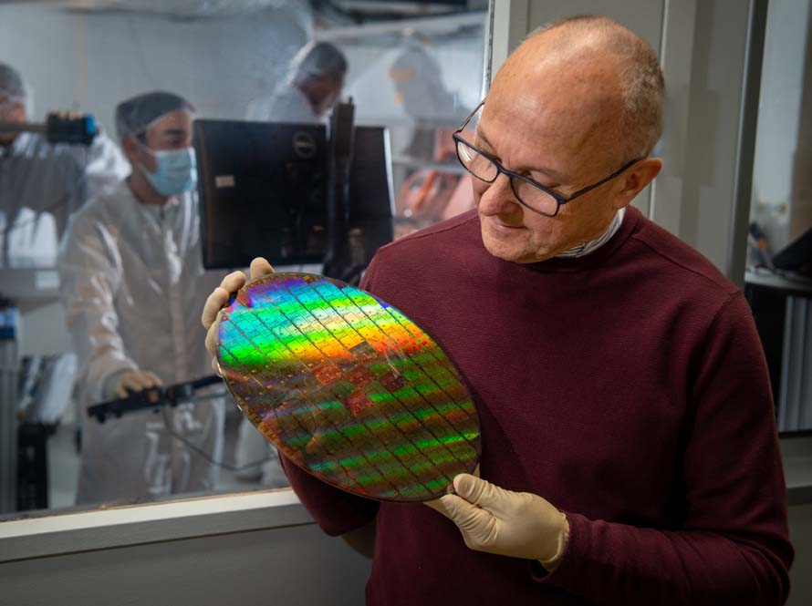 A man in a burgundy sweater holds a flat, shiny disk about the size of a dinner plate that reflects a rainbow of light across its patterned surface.