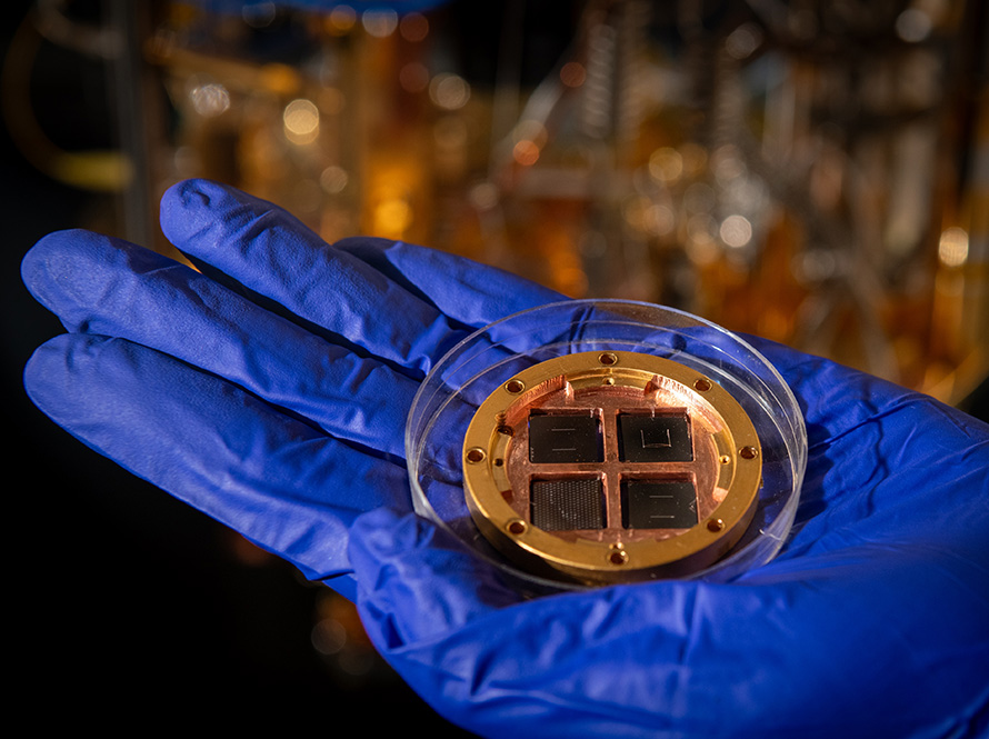 A copper colored disc-shaped device with four dark translucent panels inset lays in a clear dish, inside a blue-gloved hand.