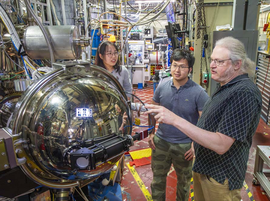 A trio of researchers stand in the ALS near a large instrument with a reflective round component that is facing the camera.