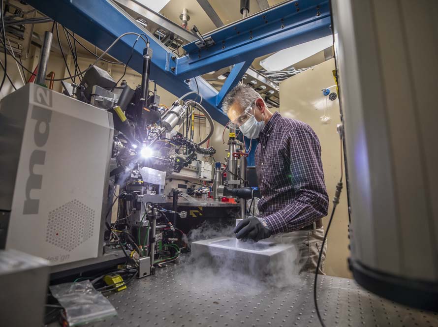 A man in a purple plaid shirt, facemask, gloves, and protective eyewear is seen adjusting a complex instrument, with sublimating liquid nitrogen used to chill samples wafting off.