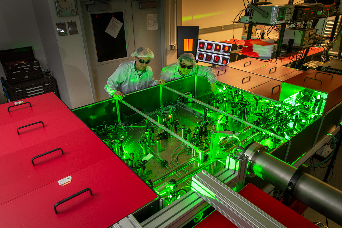 Two people wearing glasses, hair nets, and lab coats look at an array of equipment bathed in green laser light.