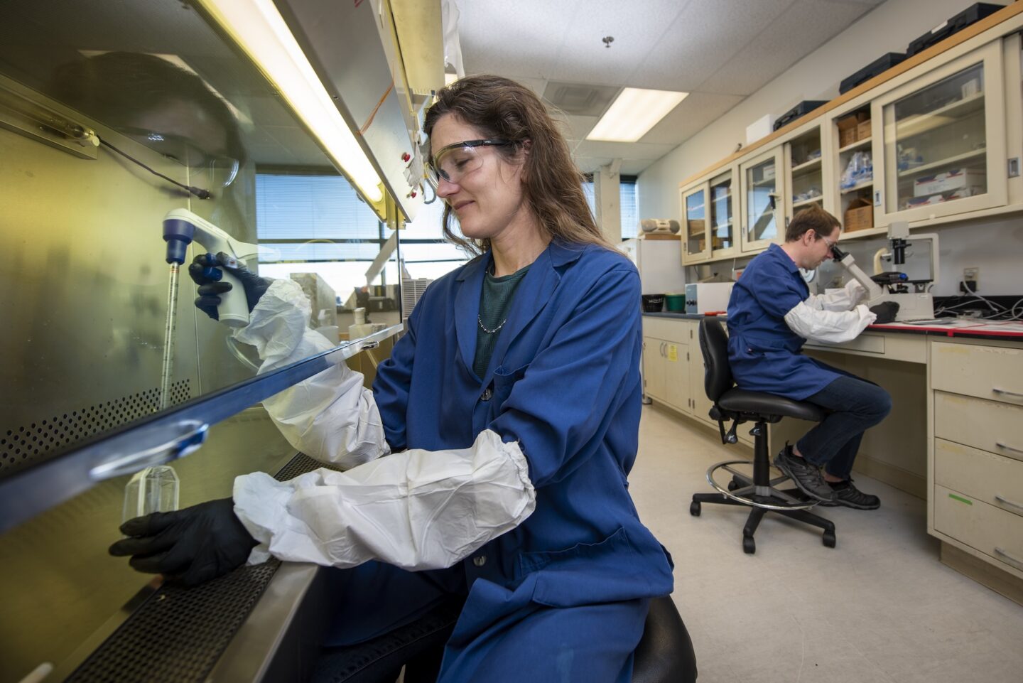 A woman in PPE pipettes liquid under a fume hood while a man works at the lab bench.